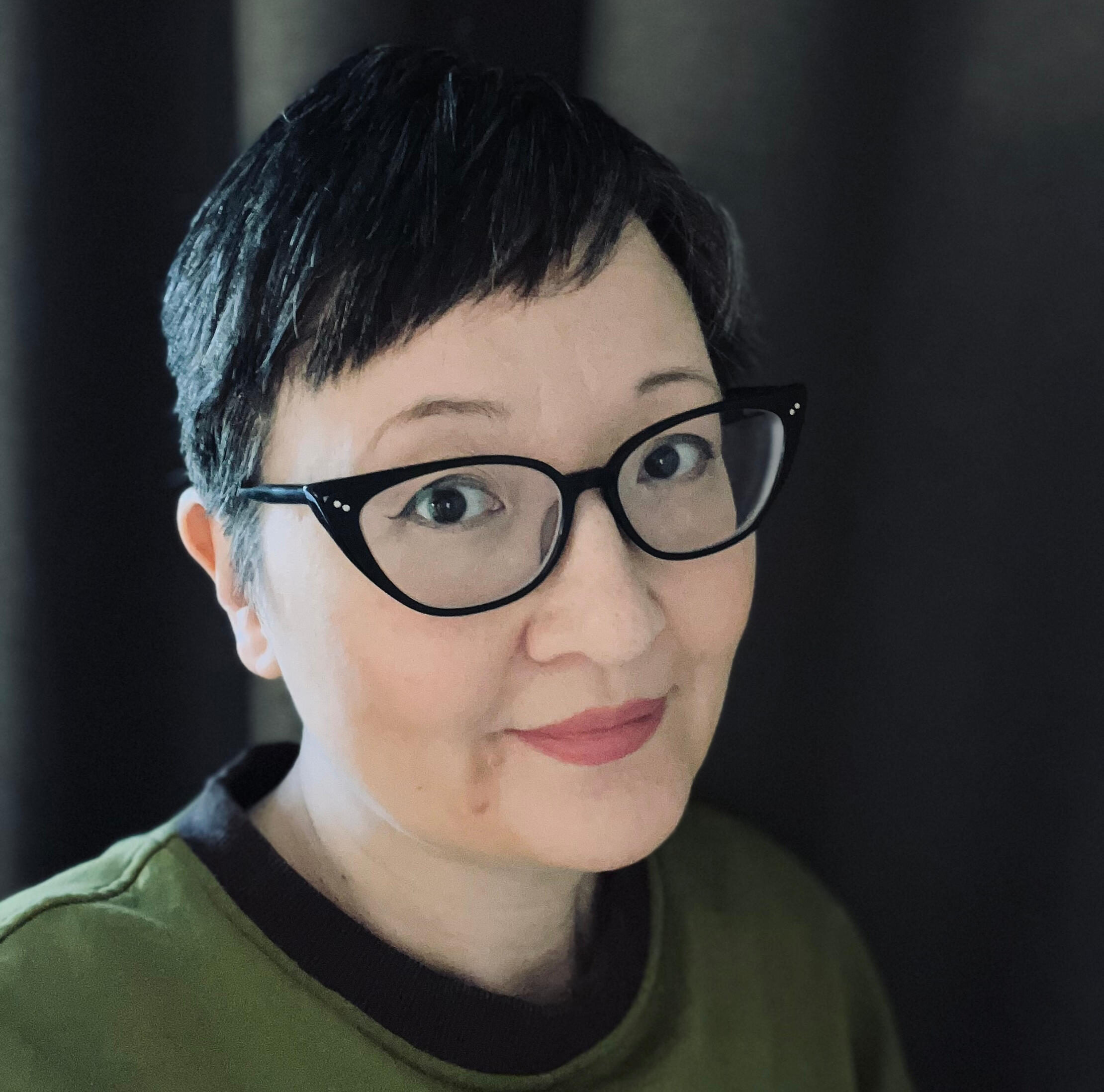 An image of me, Kat Kruger, with black hair in a pixie cut. I have black-rimmed cats eye glasses and am wearing a green top with brown trim.